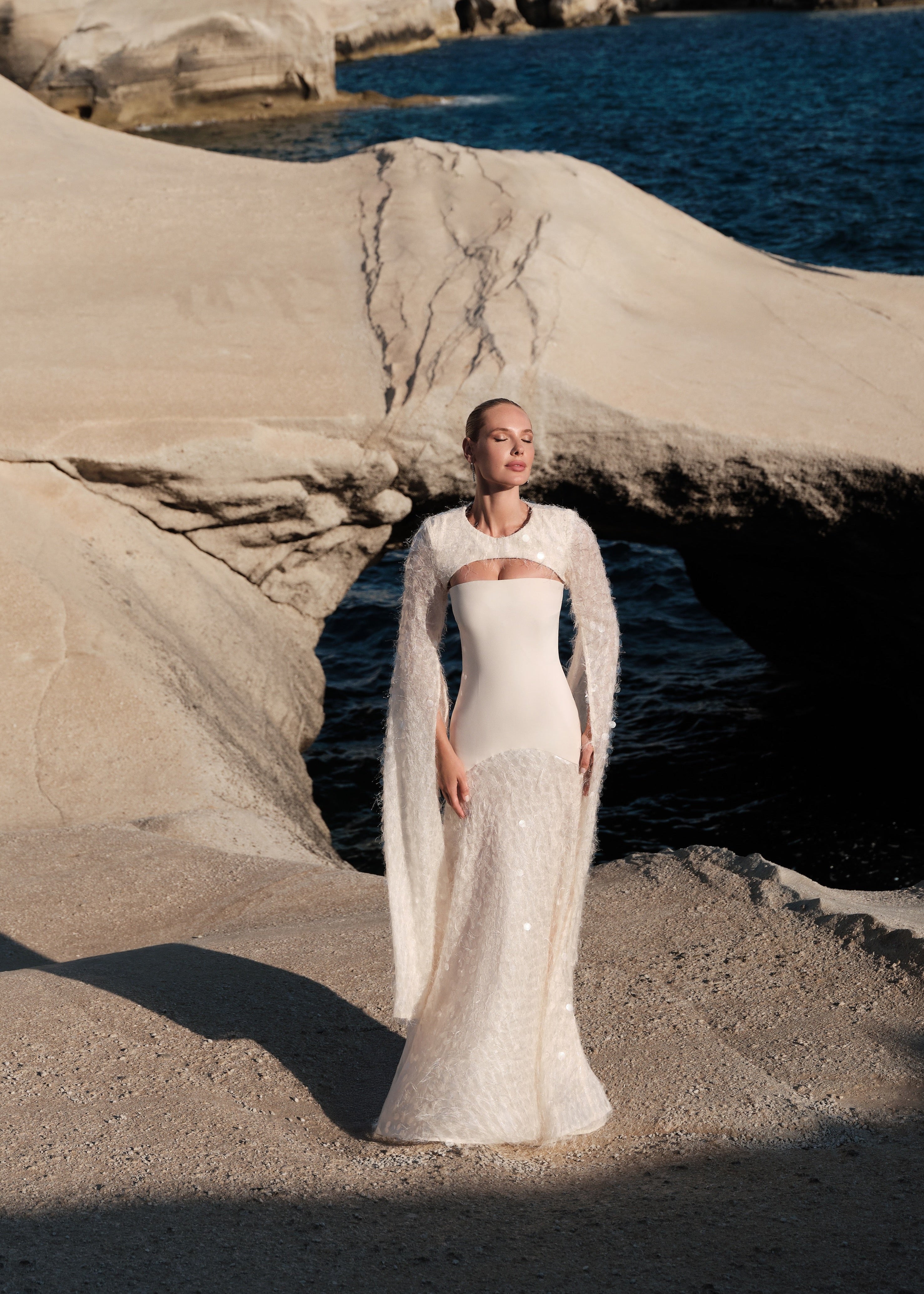 Woman in a white dress standing on a rocky beach with ocean and cliffs in the background