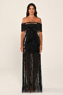 A woman modeling a black off-the-shoulder sequined maxi dress with fringe detailing on the bodice and flowing down the skirt, set against a plain light beige studio background.