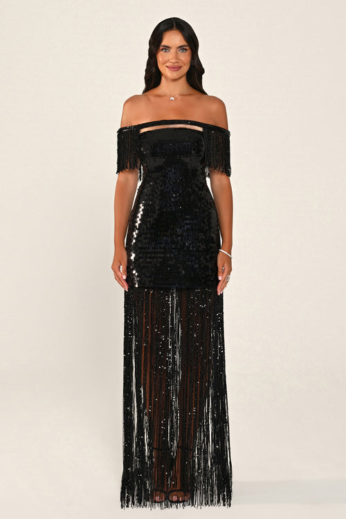 A woman modeling a black off-the-shoulder sequined maxi dress with fringe detailing on the bodice and flowing down the skirt, set against a plain light beige studio background.