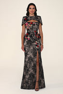 A woman modeling a floor-length black lace gown with floral applique in red, pink, and gold, featuring a high slit and structured cap sleeves, against a plain light beige studio background.