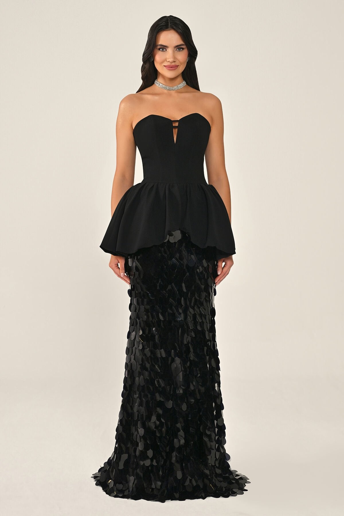 A woman modeling a strapless black gown with a corset peplum bodice and a fitted skirt of ruffled sequin petals, against a plain light beige studio background.