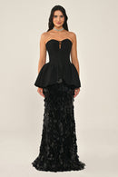 A woman modeling a strapless black gown with a corset peplum bodice and a fitted skirt of ruffled sequin petals, against a plain light beige studio background.