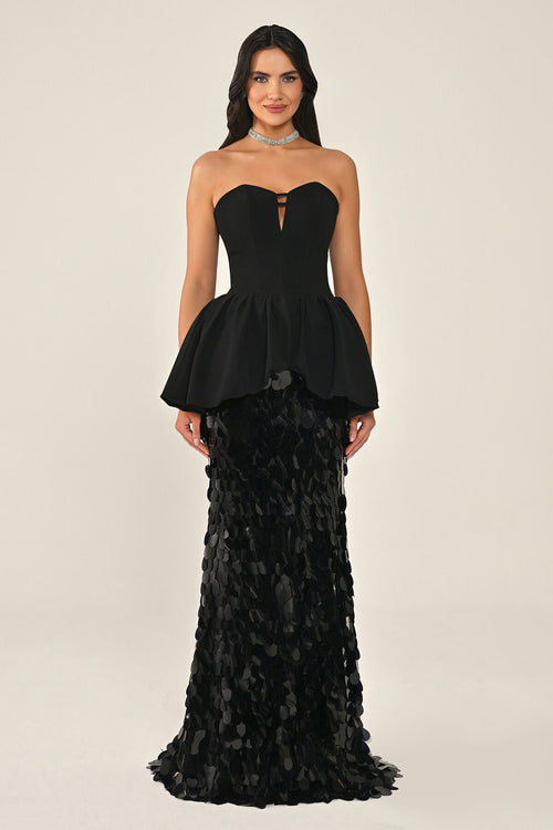 A woman modeling a strapless black gown with a corset peplum bodice and a fitted skirt of ruffled sequin petals, against a plain light beige studio background.