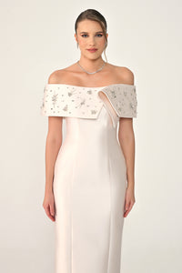 Woman wearing a white off-shoulder dress with intricate detailing on a plain background