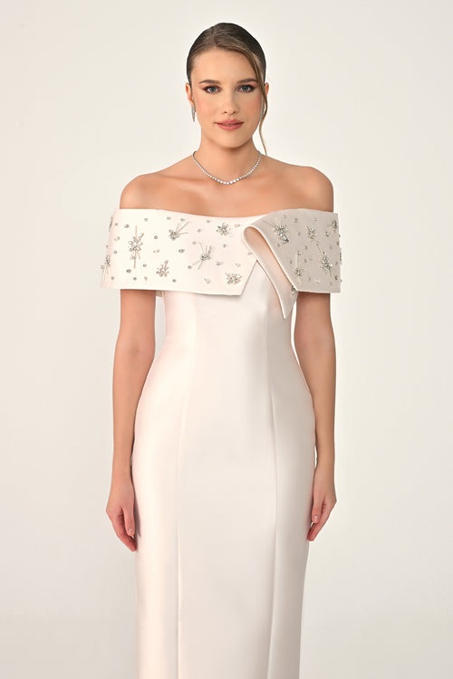Woman wearing a white off-shoulder dress with intricate detailing on a plain background