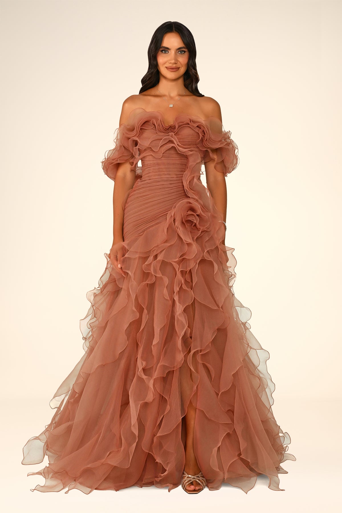 A woman modeling an off-the-shoulder, floor-length gown in a dusty rose or terracotta pink tulle fabric with dramatic ruffles and a fitted bodice, photographed against a plain warm off-white studio background.