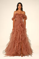 A woman modeling an off-the-shoulder, floor-length gown in a dusty rose or terracotta pink tulle fabric with dramatic ruffles and a fitted bodice, photographed against a plain warm off-white studio background.