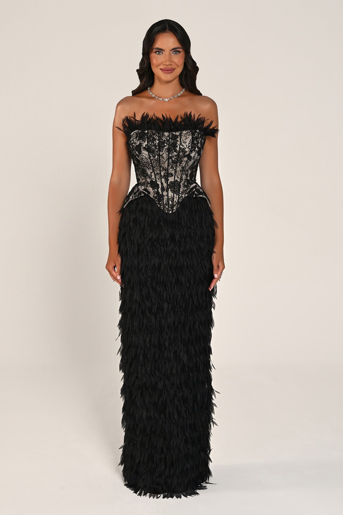 A woman modeling a strapless black evening gown with a sheer patterned corset bodice and a voluminous black feathered skirt, photographed against a plain light beige studio background.