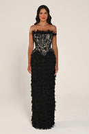 A woman modeling a strapless black evening gown with a sheer patterned corset bodice and a voluminous black feathered skirt, photographed against a plain light beige studio background.