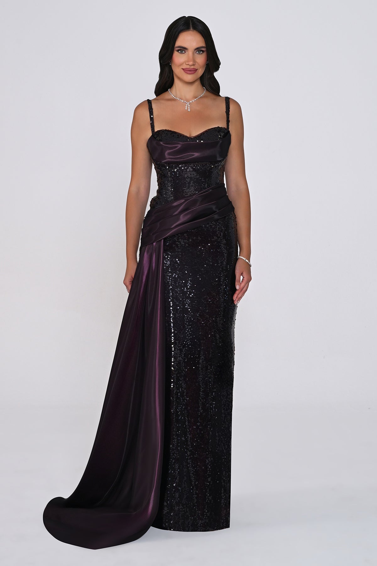 A woman modeling a floor-length evening gown with a deep plunging neckline, spaghetti straps, a sequined body, and a draped satin overskirt with a train in a dark plum color, set against a plain light gray studio background.