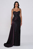 A woman modeling a floor-length evening gown with a deep plunging neckline, spaghetti straps, a sequined body, and a draped satin overskirt with a train in a dark plum color, set against a plain light gray studio background.