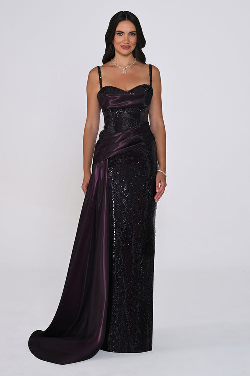 A woman modeling a floor-length evening gown with a deep plunging neckline, spaghetti straps, a sequined body, and a draped satin overskirt with a train in a dark plum color, set against a plain light gray studio background.