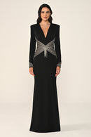 A woman modeling a floor-length black evening gown with structured shoulders, a deep V-neckline, and ornate silver beaded embellishments across the bodice and cuffs, photographed against a plain light beige studio background.
