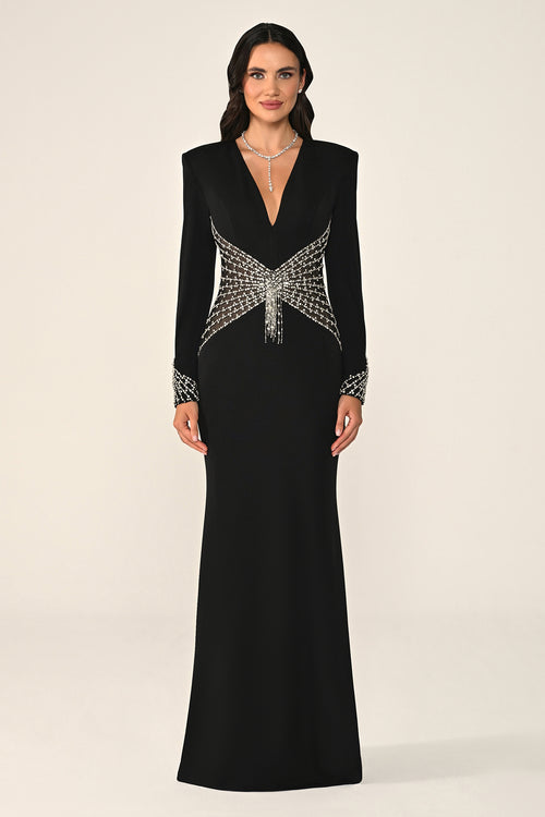 A woman modeling a floor-length black evening gown with structured shoulders, a deep V-neckline, and ornate silver beaded embellishments across the bodice and cuffs, photographed against a plain light beige studio background.