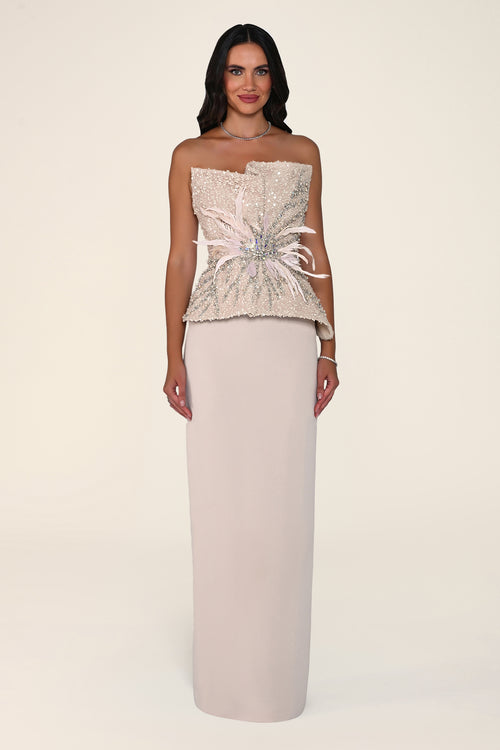 A woman modeling a strapless formal gown with a heavily embellished champagne-beige bodice featuring sequins, lace, and a large bow detail, paired with a long, solid pale blush skirt, against a solid off-white studio background.