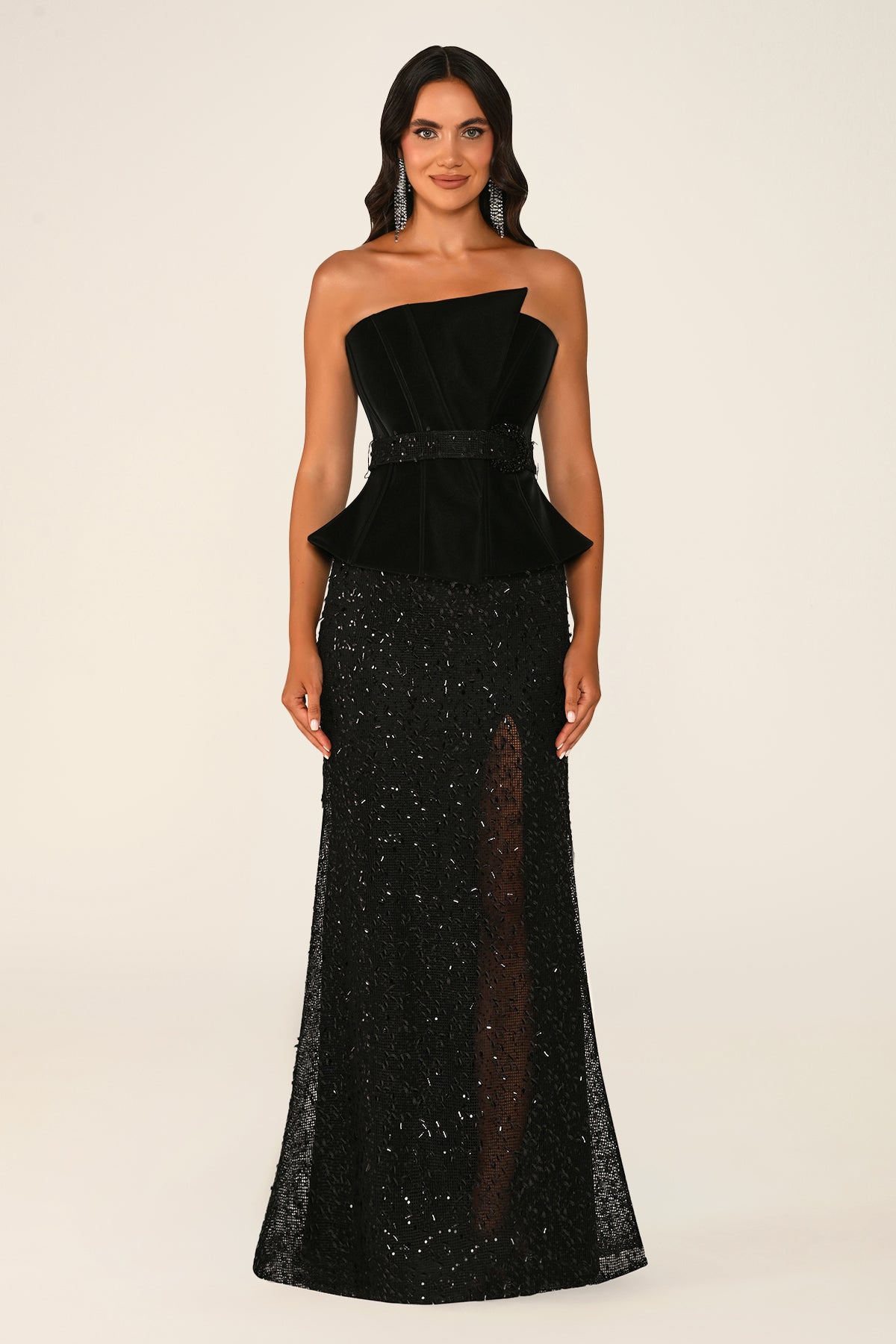 A woman modeling a black one-shoulder evening gown with a velvet peplum waist and a floor-length sequined skirt with sheer mesh detailing, photographed against a plain light beige studio background.