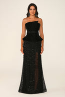 A woman modeling a black one-shoulder evening gown with a velvet peplum waist and a floor-length sequined skirt with sheer mesh detailing, photographed against a plain light beige studio background.