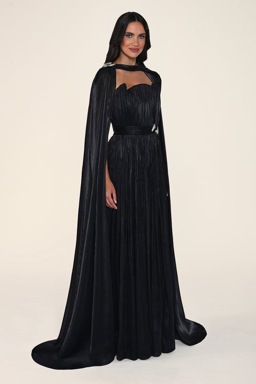 A model wearing a floor-length black pleated gown with a draped cape and a matching waist sash, photographed against a solid light beige studio background.