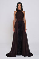 A woman modeling a floor-length dark brown halter gown with a sheer draped overlay and a central black sequined panel, photographed against a plain light gray studio background.