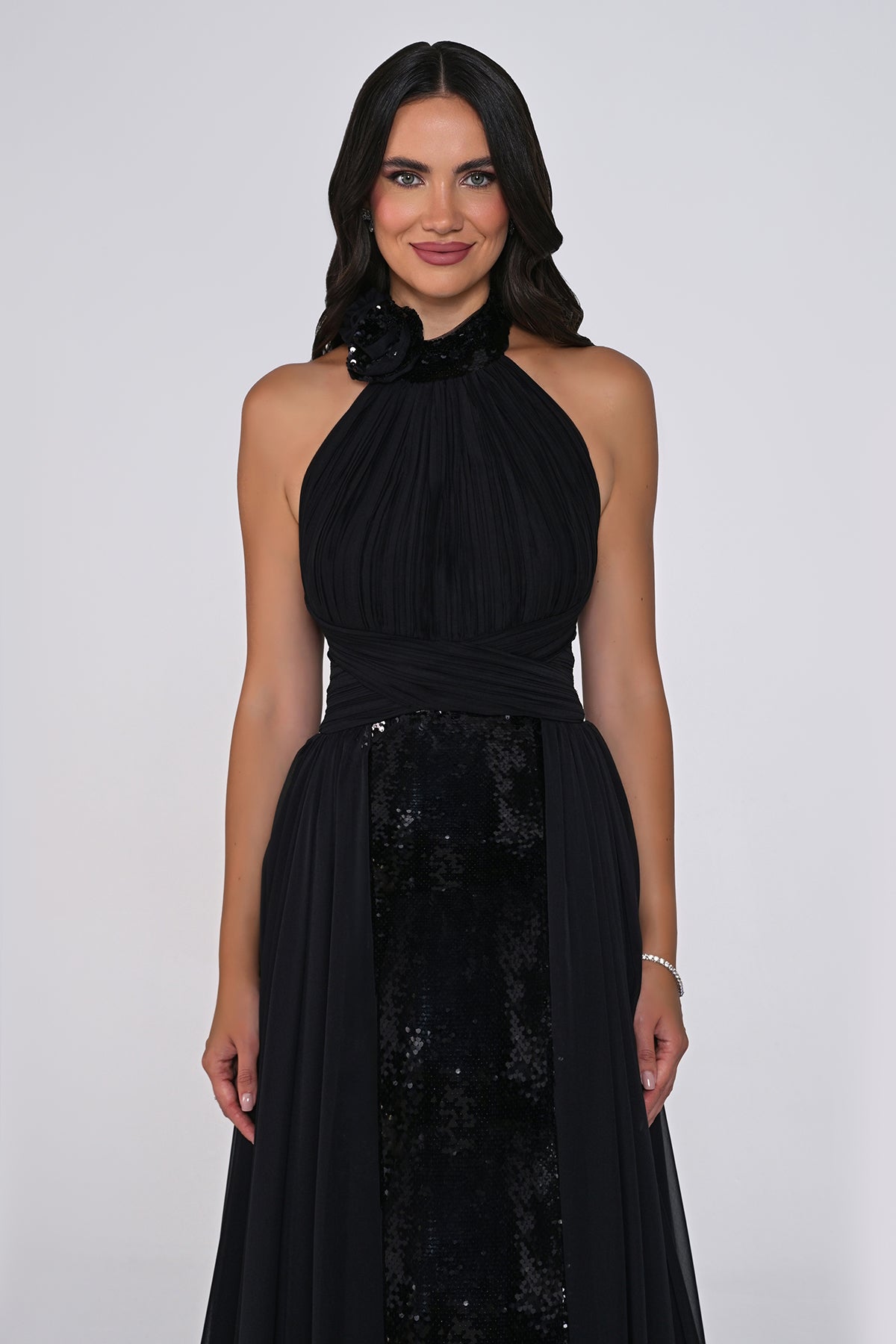 A woman modeling a sleeveless black halter-neck gown with a gathered bodice, sheer draped skirt, and a sequined panel at the front, photographed against a light gray studio background.