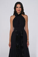 A woman modeling a sleeveless black halter-neck gown with a gathered bodice, sheer draped skirt, and a sequined panel at the front, photographed against a light gray studio background.