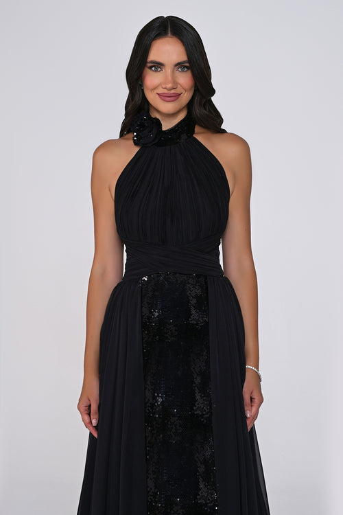 A woman modeling a sleeveless black halter-neck gown with a gathered bodice, sheer draped skirt, and a sequined panel at the front, photographed against a light gray studio background.