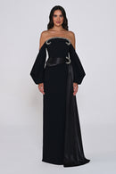 A woman modeling a floor-length black off-the-shoulder gown with dramatic balloon sleeves, a wide black waist belt, and silver embellished trim across the bodice, photographed against a light gray studio background.