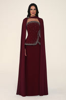 A woman modeling a floor-length burgundy gown with a matching draped cape and a heavily embellished beaded bodice, photographed against a plain light beige studio background.