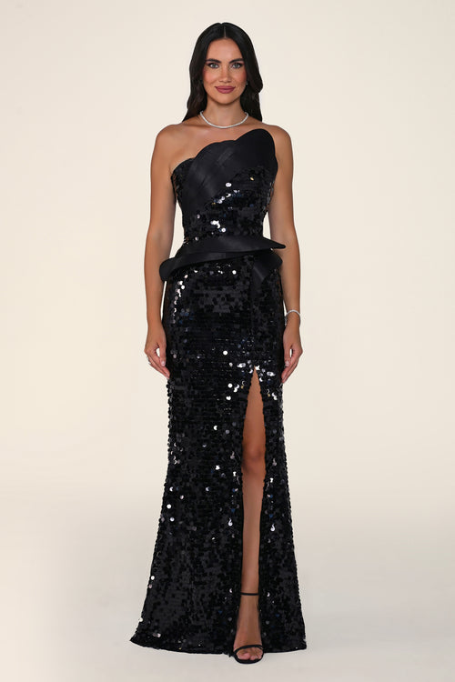 A woman modeling a floor-length black one-shoulder evening gown covered in sequins, with a high front slit and a matching black sash at the waist, against a plain light beige studio background.