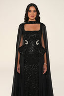 Celestara Dress in Black: sequin column gown with a square neckline, peplum hip detail, stone-embellished waist and a flowing organza cape