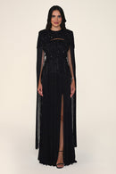 A model wearing a long black evening gown with sheer long sleeves, a beaded bodice, and a high thigh slit, photographed against a plain light beige studio background.