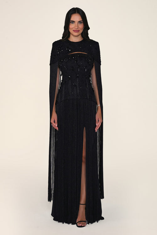 A model wearing a long black evening gown with sheer long sleeves, a beaded bodice, and a high thigh slit, photographed against a plain light beige studio background.