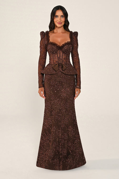 A woman modeling a floor-length brown lace gown with long puff sleeves, a sweetheart neckline, a lace-up corset-style bodice, and a peplum waist, photographed against a plain light beige studio background.
