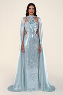 A woman modeling a floor-length light blue sequined gown with a matching sheer cape, floral embellishment at the neckline, and side cutouts, photographed against a plain warm beige studio background.