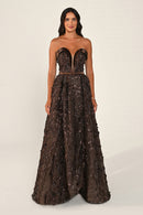 A woman modeling a strapless floor-length gown with a sweetheart neckline, fitted corset-style bodice, and a voluminous heavily embellished skirt in dark brown and bronze tones, photographed against a plain light beige studio background.
