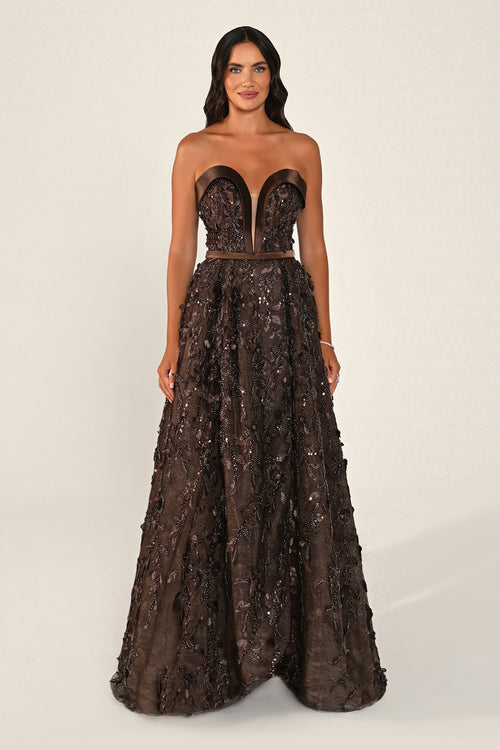 A woman modeling a strapless floor-length gown with a sweetheart neckline, fitted corset-style bodice, and a voluminous heavily embellished skirt in dark brown and bronze tones, photographed against a plain light beige studio background.