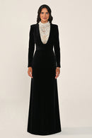 A woman modeling a floor-length black velvet gown with long sleeves and a structured collar, paired with a layered cream beaded necklace, against a plain light beige studio background.