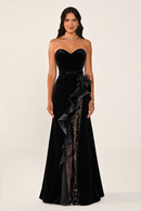 A woman modeling a strapless black velvet evening gown with a sweetheart neckline, a fitted waist, and dramatic layered ruffle draping over one side of the skirt, photographed against a plain light beige studio background.