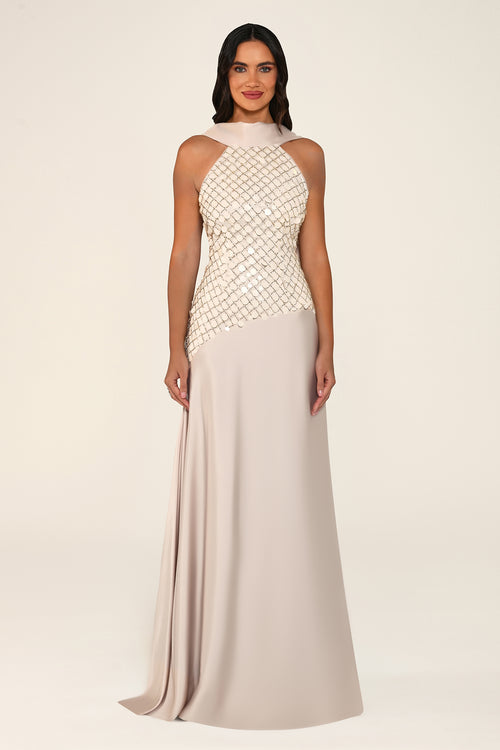 A woman modeling a sleeveless floor-length gown with a high mock neck, a shimmering beaded bodice, and a flowing satin skirt in a light champagne and blush beige tone, set against a plain cream studio background.