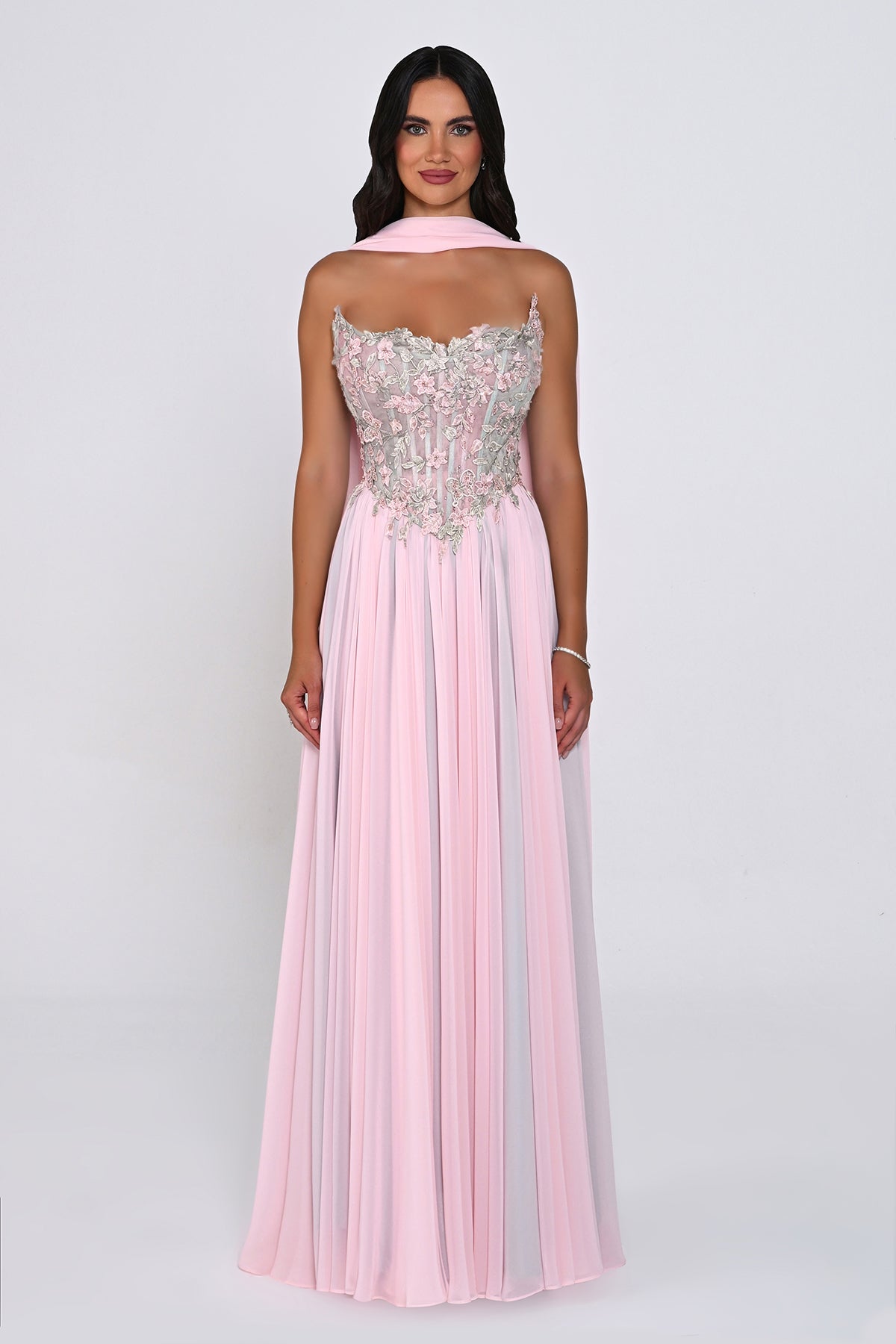 A woman modeling a floor-length formal gown from the back against a light gray studio background. The dress has a pale pink pleated skirt, a matching pink halter-style upper section, and a heavily embellished bodice with silver and pink floral embroidery.