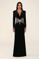 A woman modeling a floor-length black velvet gown with a plunging V-neckline, long sleeves, and a wide embellished waistband with silver rhinestone-like detailing, against a plain light beige studio background.