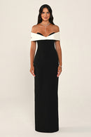 A woman modeling a floor-length off-the-shoulder gown with a black body and contrasting white shoulder panels, photographed against a plain light beige studio background.