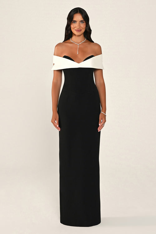 A woman modeling a floor-length off-the-shoulder gown with a black body and contrasting white shoulder panels, photographed against a plain light beige studio background.