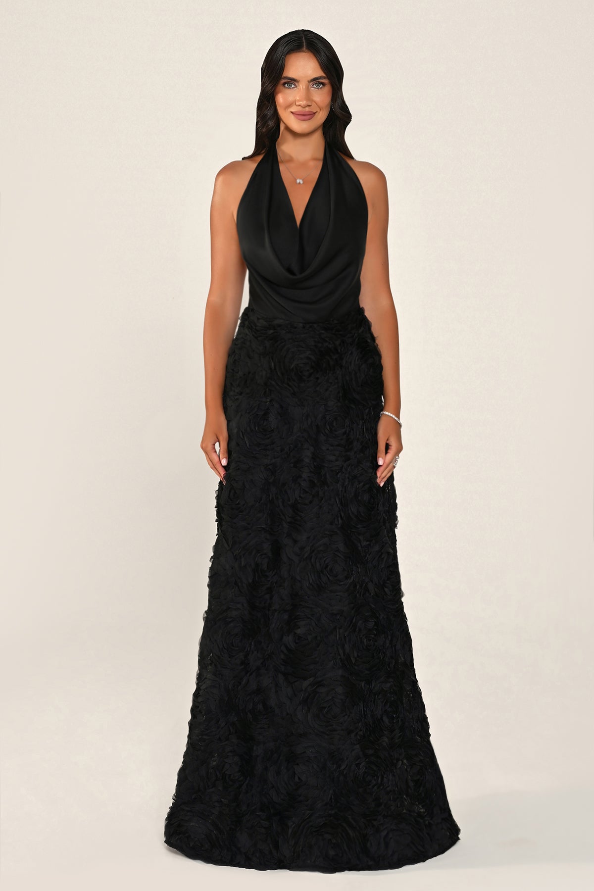 A woman modeling a sleeveless black halter-neck maxi dress with a draped cowl neckline and a textured, ruffled floor-length skirt, photographed against a plain light beige studio background.