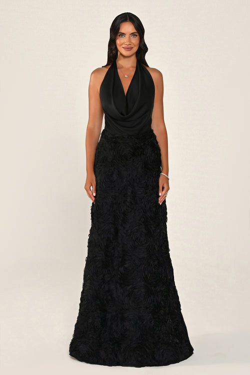 A woman modeling a sleeveless black halter-neck maxi dress with a draped cowl neckline and a textured, ruffled floor-length skirt, photographed against a plain light beige studio background.