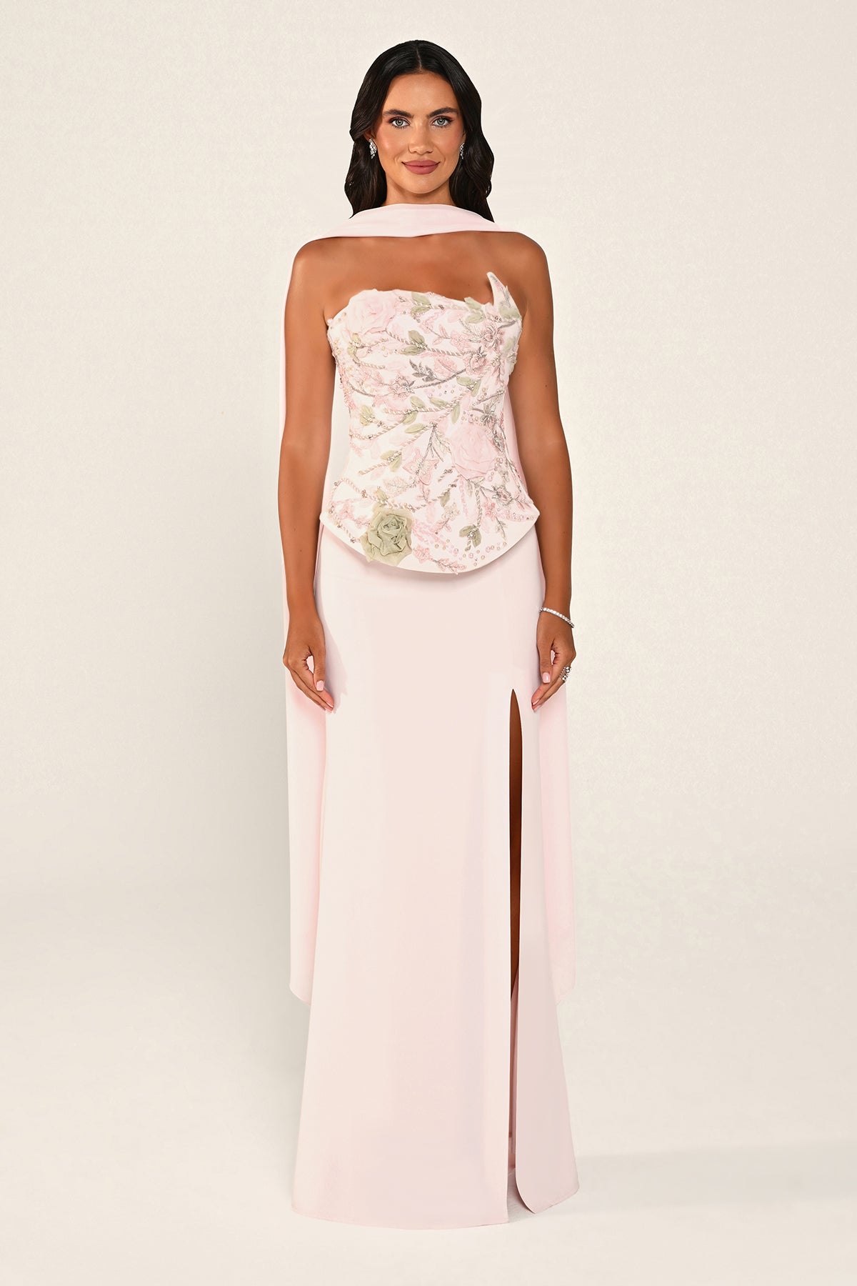 A woman modeling a pale pink strapless gown with a floral embroidered bodice and a high front slit, photographed against a plain light beige studio background.