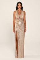 A woman modeling a floor-length gold sequin gown with a draped cowl neckline, halter straps and a high side slit, against a plain light beige studio background.