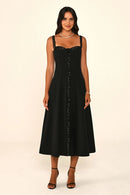 A woman modeling a sleeveless black midi dress with thin straps, a lace-trimmed sweetheart neckline, and a vertical line of embellished buttons down the front, set against a plain cream studio background.