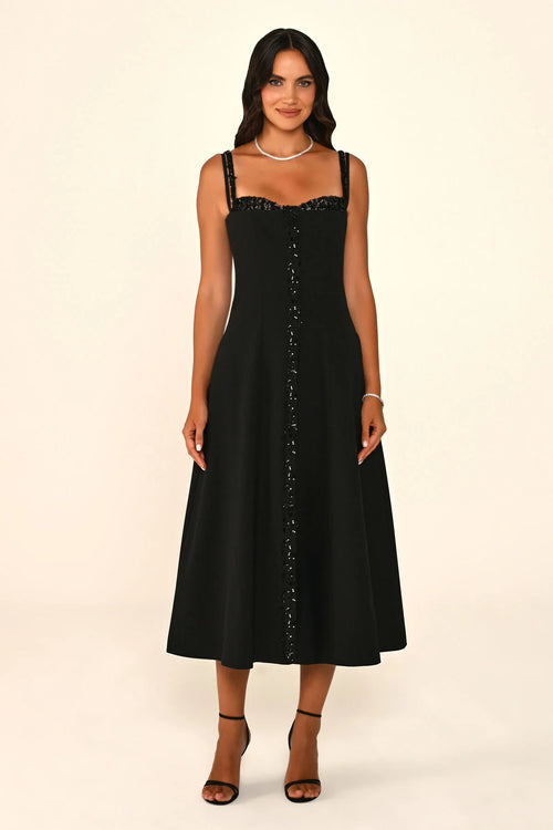 A woman modeling a sleeveless black midi dress with thin straps, a lace-trimmed sweetheart neckline, and a vertical line of embellished buttons down the front, set against a plain cream studio background.