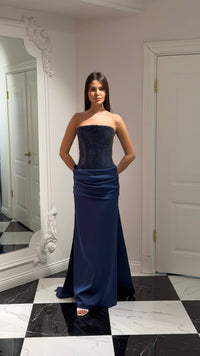 Woman wearing a strapless navy blue gown standing in a room with a mirror and white walls.